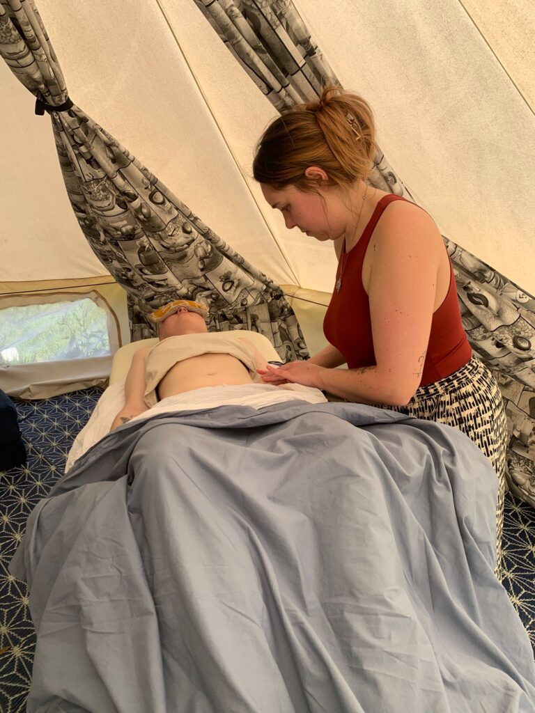 Ram & Rose Alchemy is pictured to the right of a client who is experiencing Integrative Womb Hara Massage. The client is laying on a massage bed, draped in cosy materials and wears an eye mask. 
