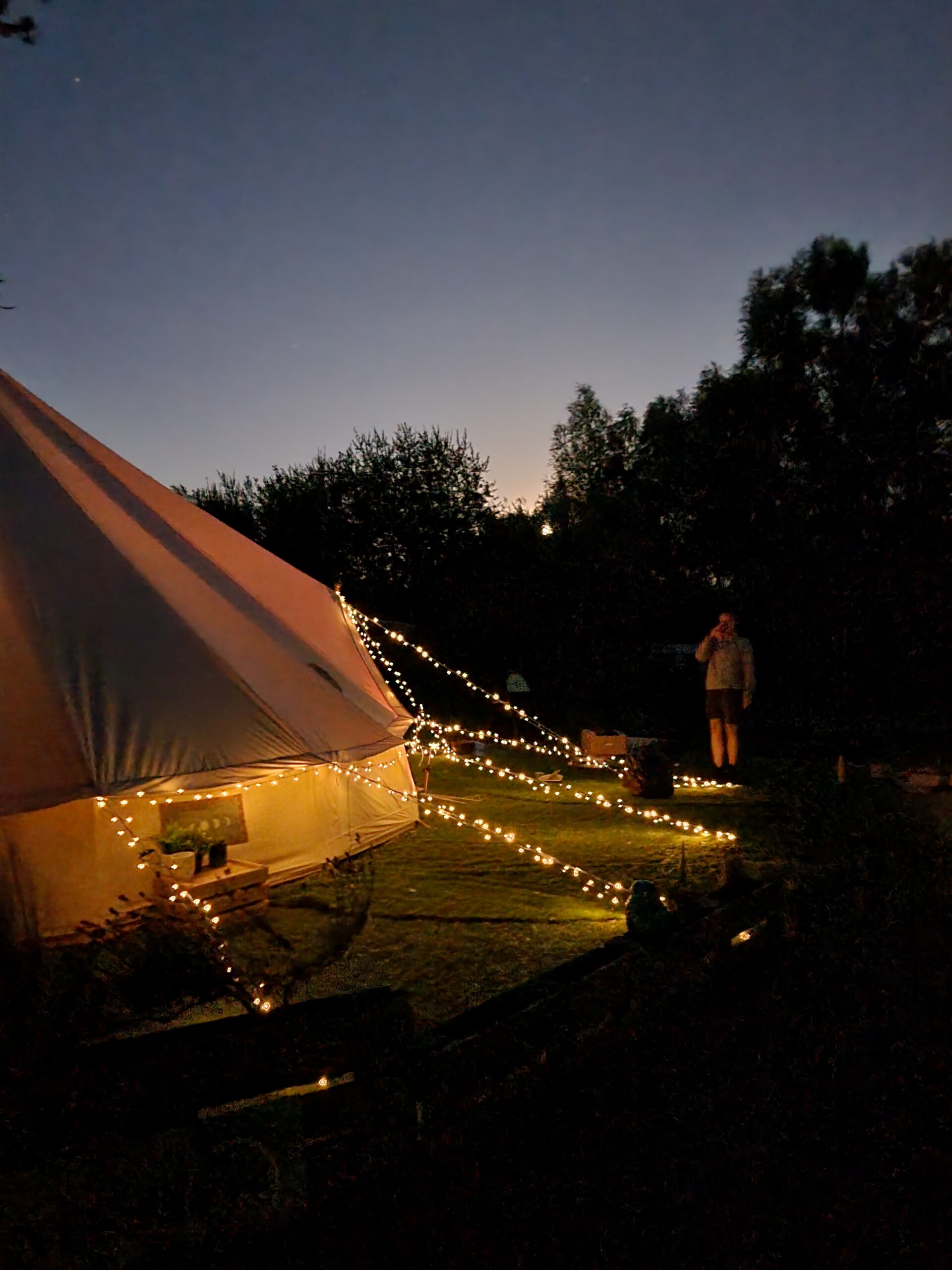 A dusky sky sets the evening scene over The Dragonfly Hideaway. Fairly lights adorn the bell tent and create an amber glow.